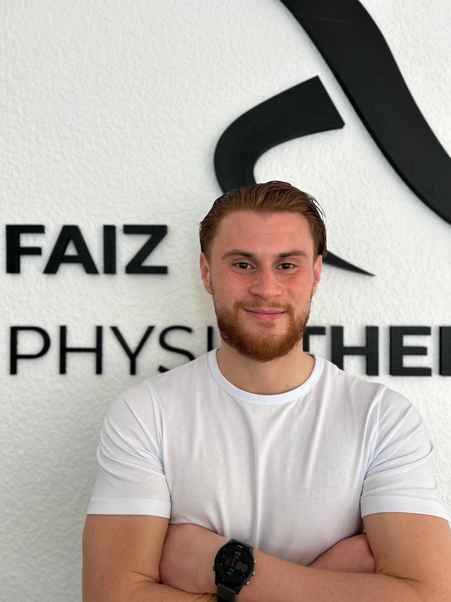Mohammed Faiz, Gründer von Faiz Physiotherapie, steht lächelnd vor der Praxiswand mit FAIZ und PHYSIO Beschriftung